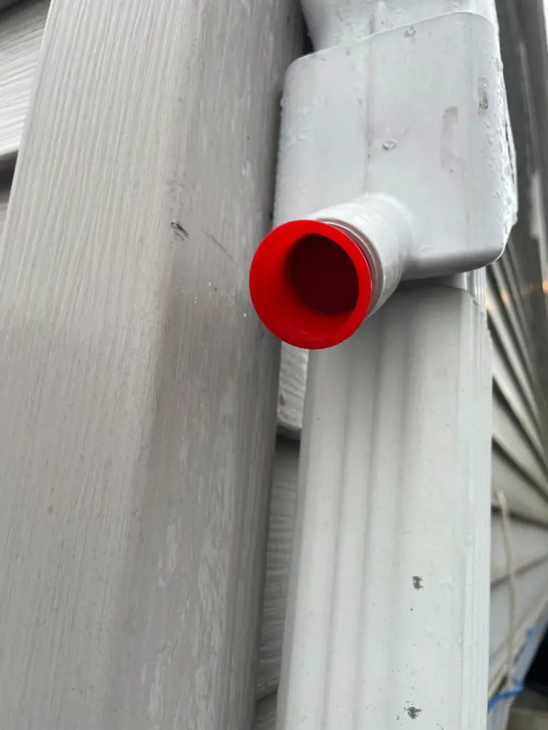 A rainwater collecting gutter kit attached to the gutter of a house. There is a red stopper to keep the water flowing through the gutter.